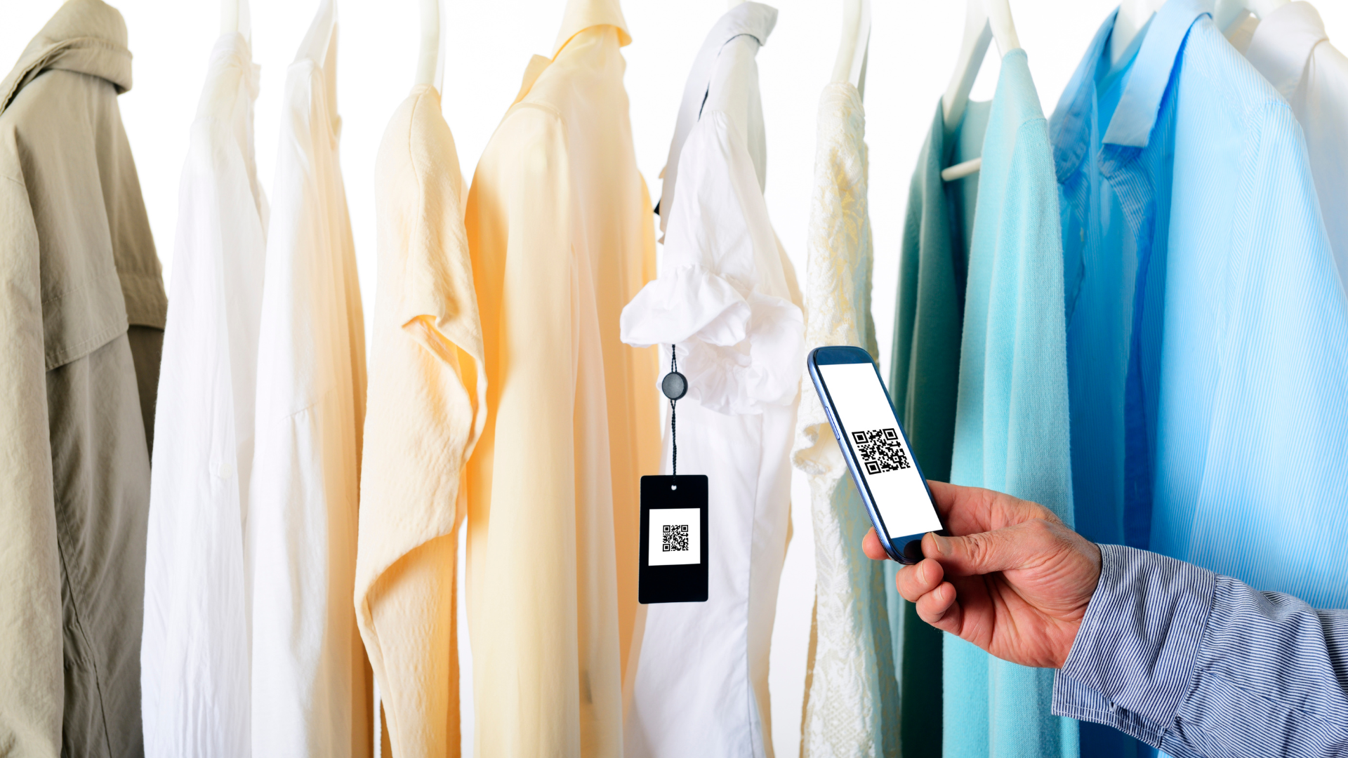 A person scans a QR code hanging on a white shirt among various pastel-colored clothes on hangers using a smartphone.