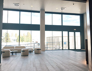 Spacious lobby with large windows, white walls, wood floor, and minimal seating. Natural light floods the space.
