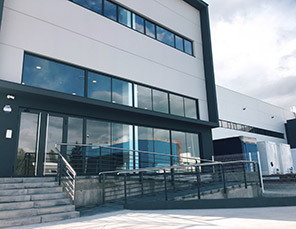Modern industrial building with large windows, a glass entrance, and an adjacent ramp. The sky is partly cloudy.
