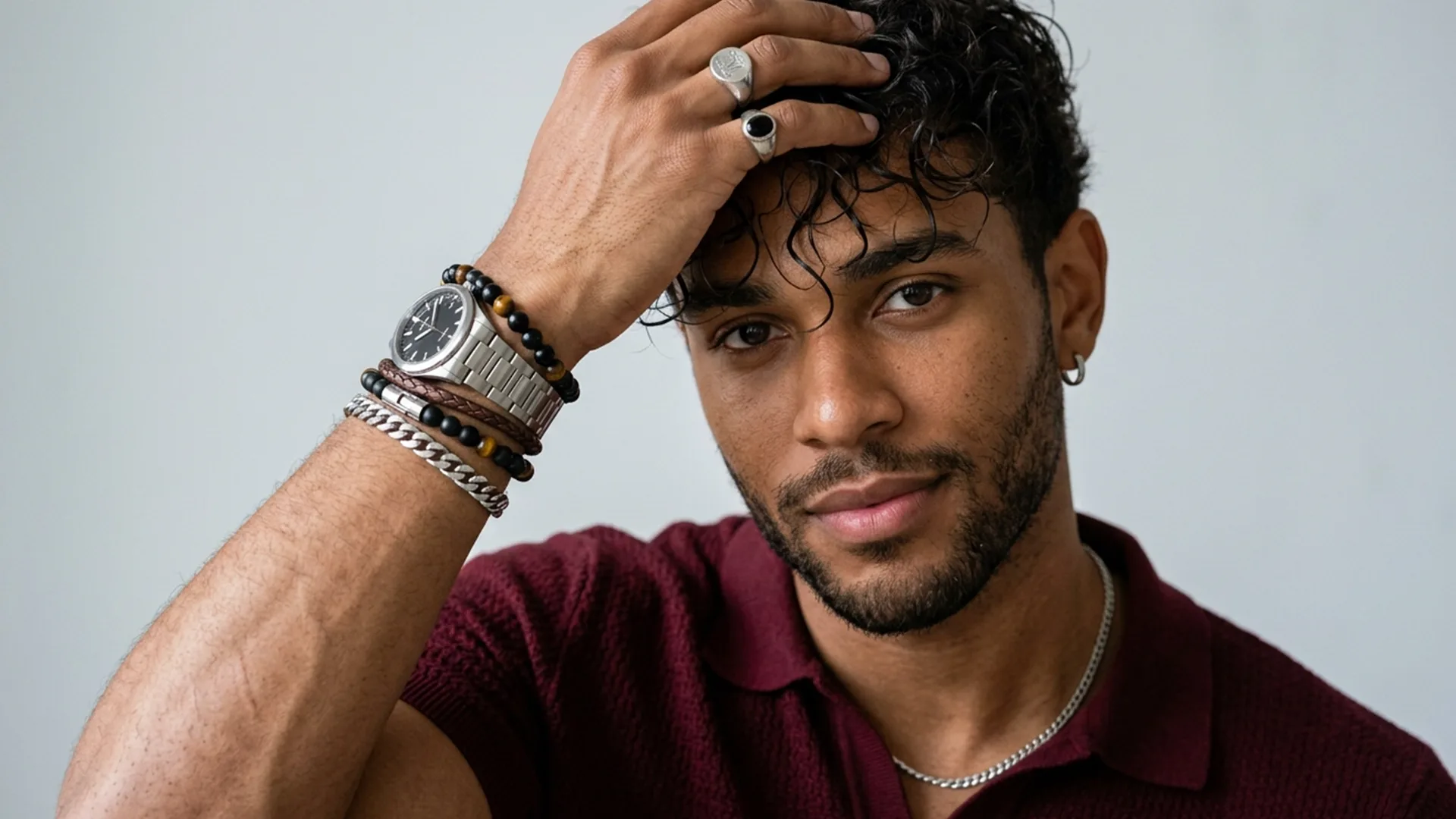 Man with short curly hair wearing a maroon shirt, bracelets, rings, and a watch, touching his head and looking at the camera against a plain background.