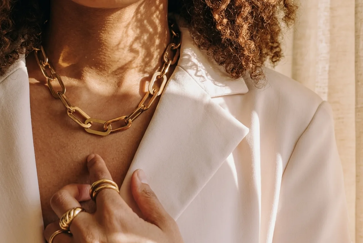 A person wearing a white blazer, a chunky gold chain necklace, and gold rings touches their chest.
