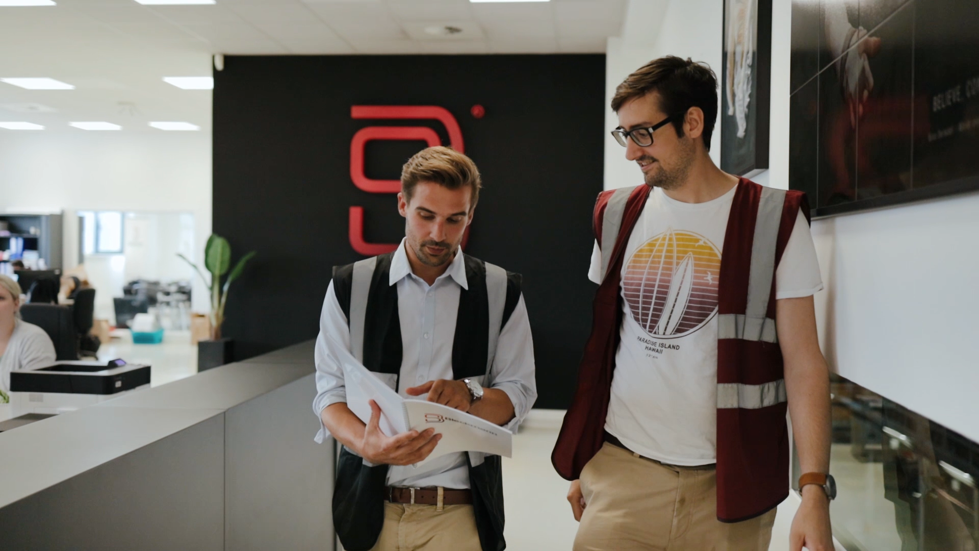 Two men wearing safety vests walk indoors, reviewing documents together near a black wall with a red logo.