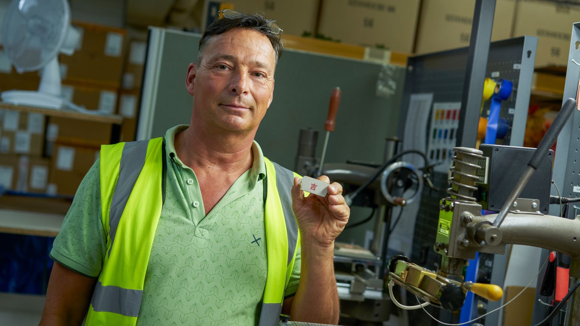 Man in a green polo and reflective vest holds a small object in an industrial workspace, surrounded by machinery and boxes.