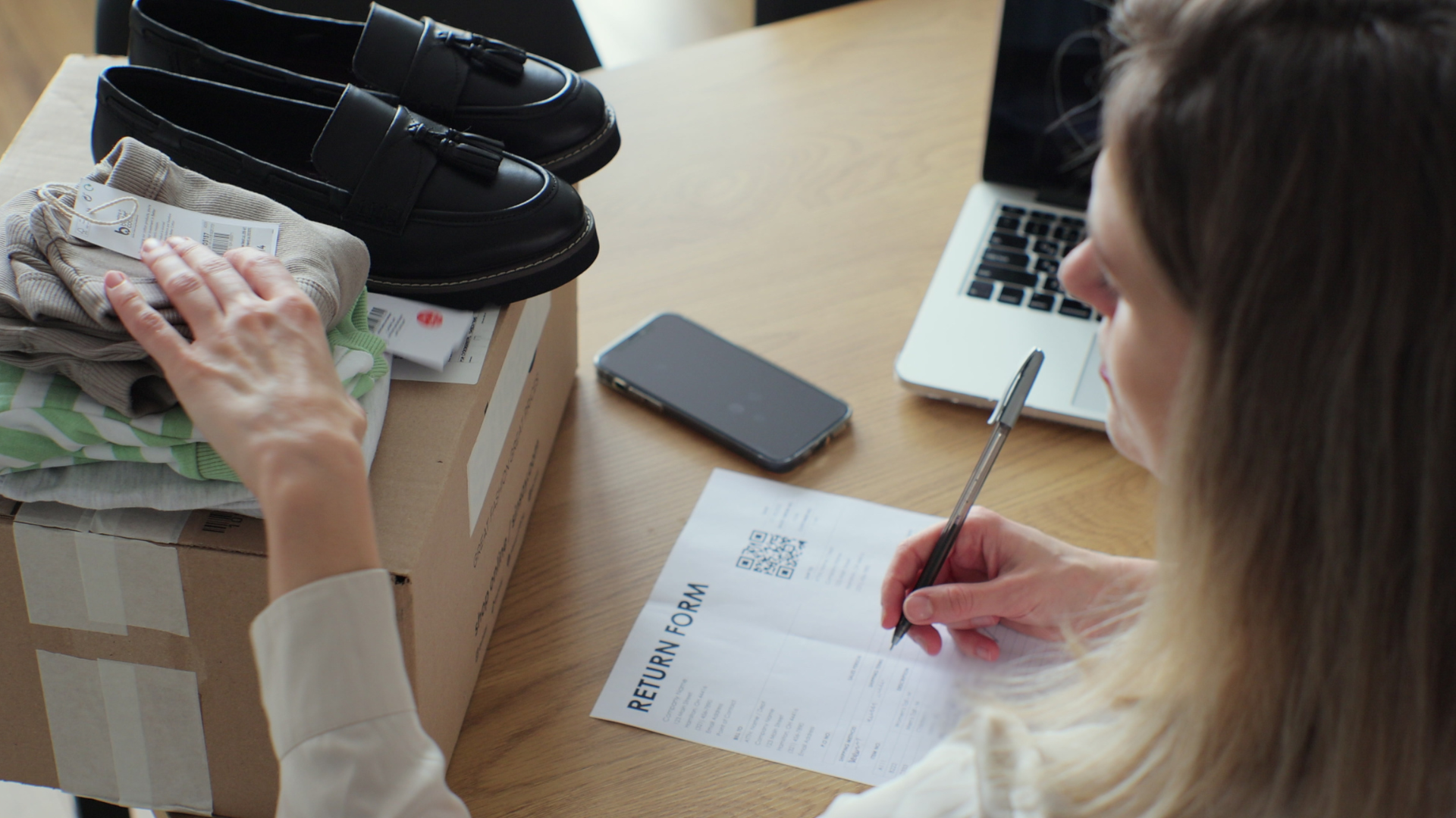 A person fills out a return form next to a box with clothes and shoes, a smartphone, and a laptop on a wooden table.