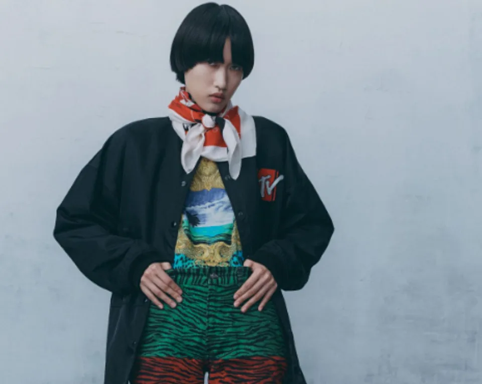 Person with short black hair wearing a black jacket, colorful patterned shorts, and a red and white scarf, standing against a light-colored wall.