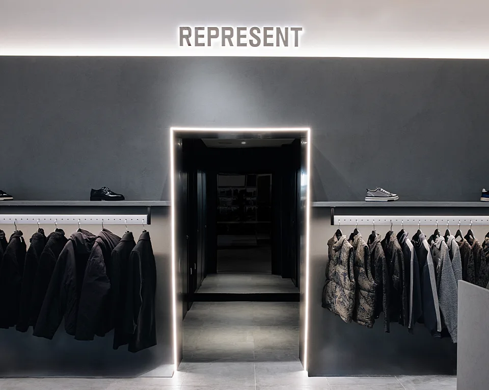 Clothing display in a modern store featuring jackets and shoes. A central, illuminated doorway leads to another area, with "REPRESENT" signage above.