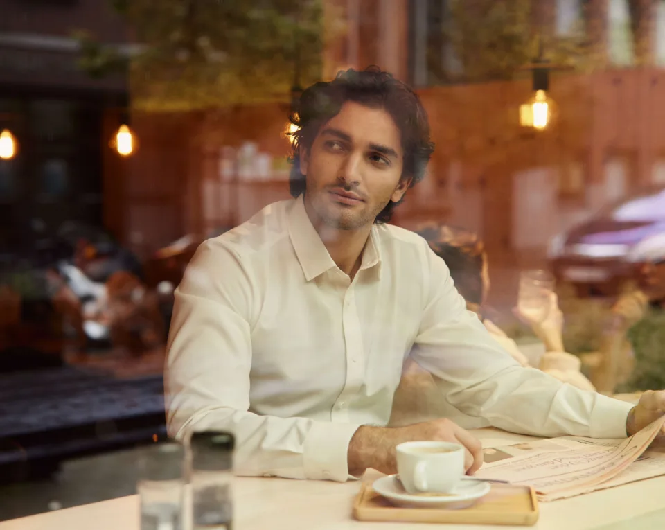 A person in a white shirt is sitting at a cafe table, holding a newspaper. A cup of coffee is in front of them. Reflections of street and lights are visible on the window.
