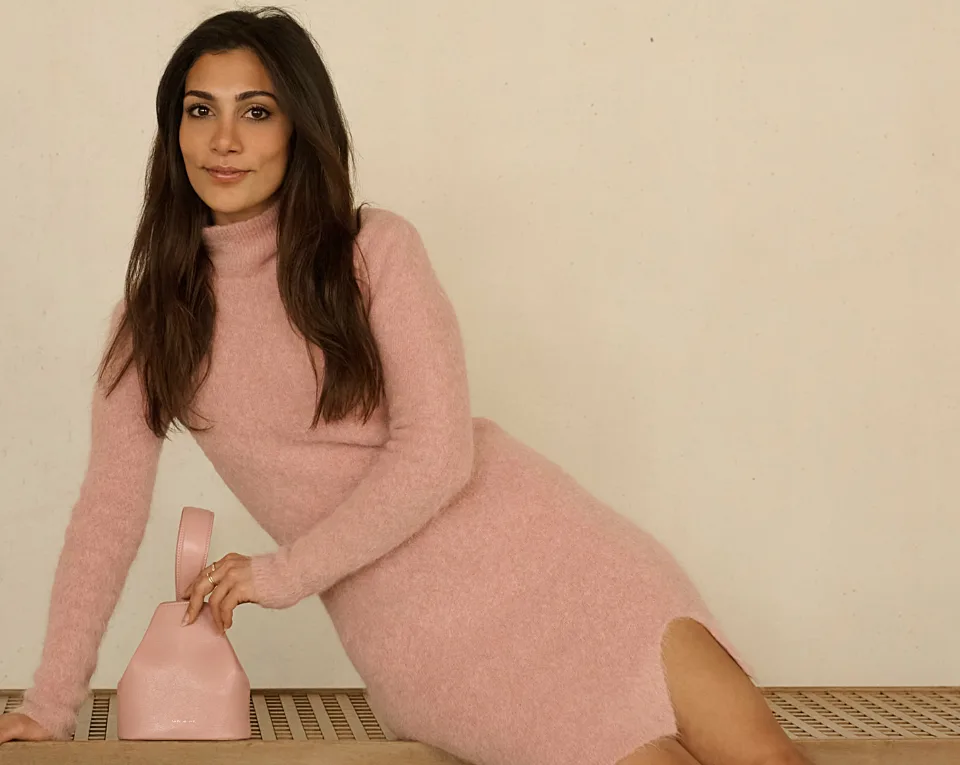 A woman in a light pink long-sleeve dress sits on a bench, with her hand resting on a small pink handbag against a plain background.