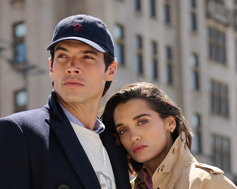 A man in a dark cap and coat stands beside a woman in a beige jacket, both looking into the distance with a city building in the background.