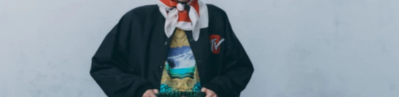 Person with short black hair wearing a black jacket, colorful patterned shirt, red and green shorts, and a white and red scarf, standing against a light gray wall.