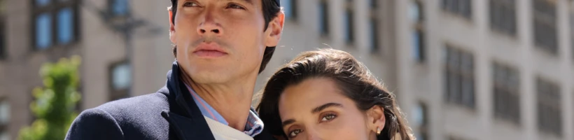 A man in a navy cap and coat stands next to a woman in a beige trench coat, with buildings in the background.