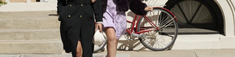 Two women walk and laugh on a city sidewalk; one wears a black coat, the other a purple dress, and a red bicycle is parked behind them near concrete steps.