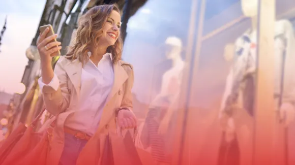 Woman walking with shopping bags and holding a smartphone, smiling while passing by store windows displaying mannequins.