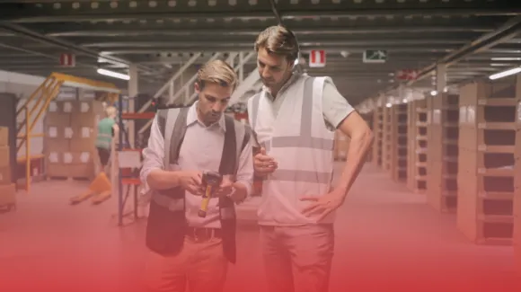 Two men stand in a warehouse, one holding a handheld scanner. They are surrounded by shelves filled with boxes.