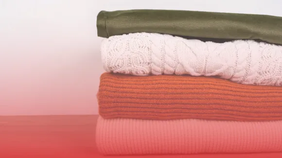 A neatly stacked pile of three sweaters in textured patterns and solid colors, placed on a pink surface against a white background.