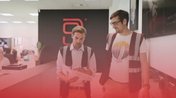 Two men wearing vests discuss a document in an office with a modern design and a logo on the wall.