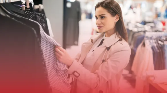 A woman in a trench coat examines clothing on a rack in a store.