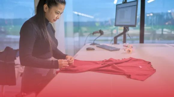 A woman works at a desk, examining a red garment. A computer with a monitor is in the background.