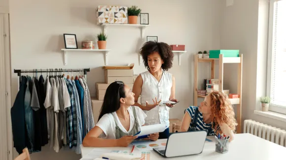 Three women engage in discussion around a desk with a laptop. Clothing on hangers and shelves with decor are in the background.