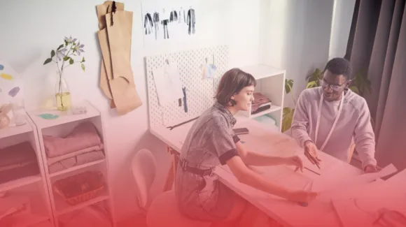 Two people in a design studio with sewing patterns and fabric samples on a table, surrounded by design sketches and materials.