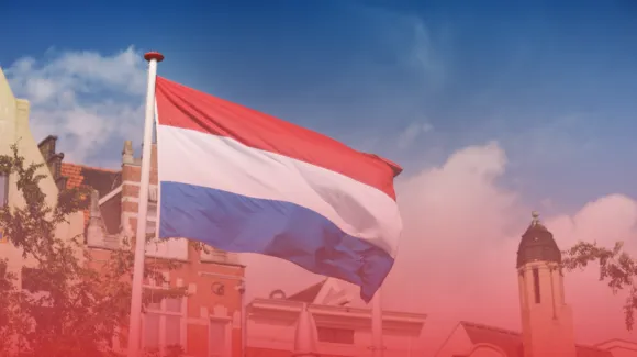 The Dutch flag waves against a clear blue sky with historical buildings in the background.