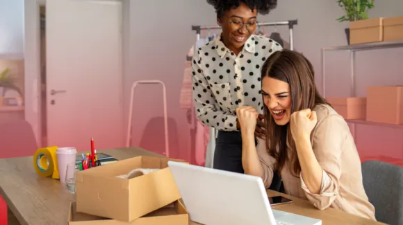 Two women in an office celebrate while looking at a laptop, with shipping boxes, tape, and office supplies on the table around them.