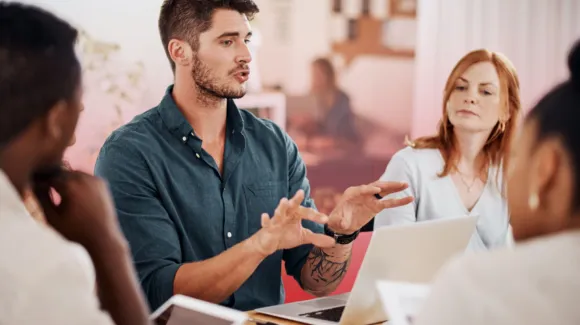 A man speaks while gesturing with his hands during a meeting with three colleagues, seated around a table with laptops and documents.