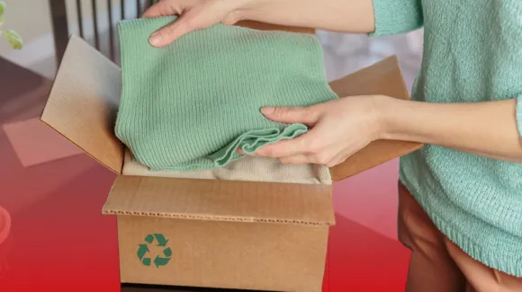 A person folds a green sweater into a cardboard box with a recycling symbol on it, suggesting packaging or donation.