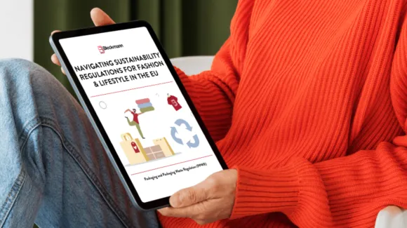 A person in an orange sweater holds a tablet displaying a document titled "Navigating Sustainability Regulations for Fashion & Lifestyle in the EU.