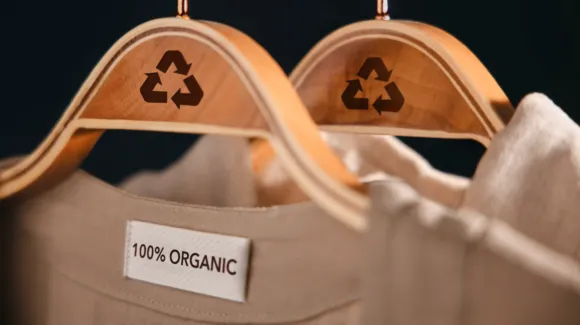 Two wooden hangers with recycling symbols hold beige clothing, one labeled "100% ORGANIC," against a dark background.