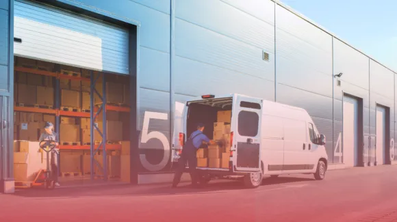 Two workers load cardboard boxes from a warehouse into a white delivery van parked outside large industrial storage units.