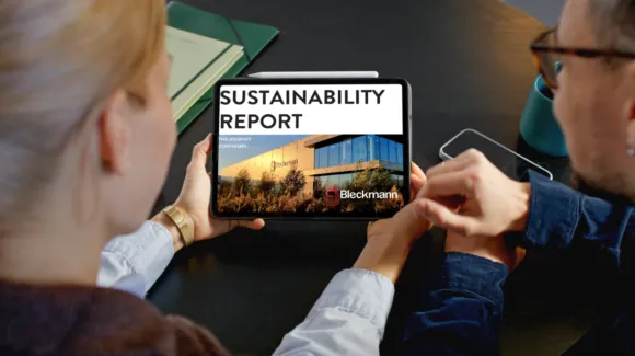 Two people view a tablet displaying a sustainability report with the title “SUSTAINABILITY REPORT” and the Bleckmann logo on the cover, seated at a table with documents and a smartphone.