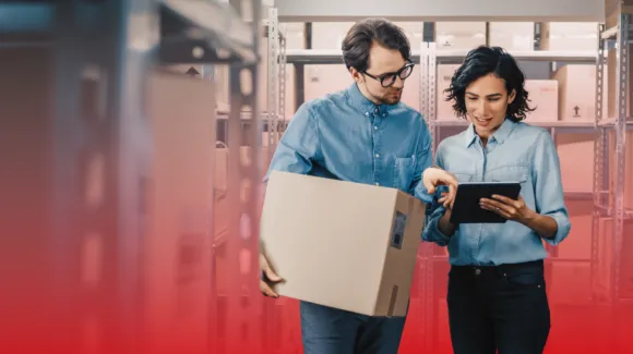 Two people standing in a storage room; one holds a cardboard box while the other points at a tablet, both appear to be discussing inventory.