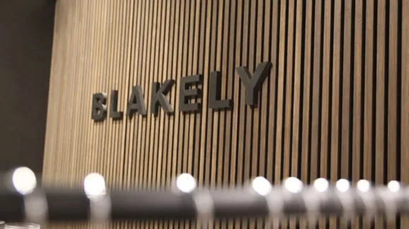 Black letters spelling "BLAKELY" mounted on a wall with vertical wooden slats, with a blurred coat rack in the foreground.