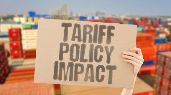 A person holds a cardboard sign that reads "TARIFF POLICY IMPACT" with shipping containers and an industrial port in the background.