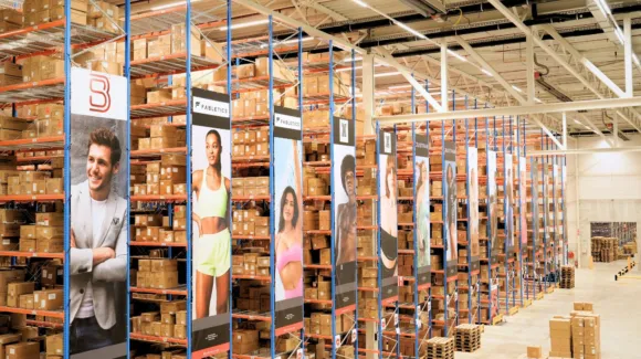 Large warehouse with tall shelves filled with boxes; banners featuring people in athletic or casual wear hang from shelving units.