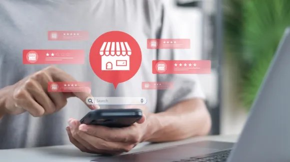 A person uses a smartphone, with a laptop nearby, as digital icons of store fronts and review ratings appear, representing online business search and customer reviews.