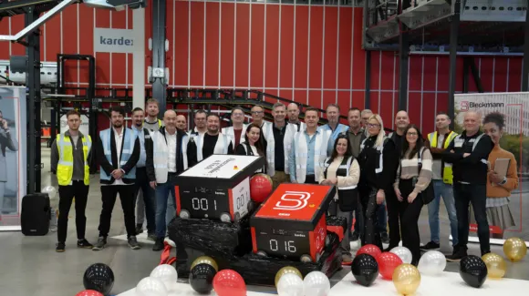 A group of people poses indoors with two large numbered boxes and scattered balloons, in an industrial setting with machinery in the background.