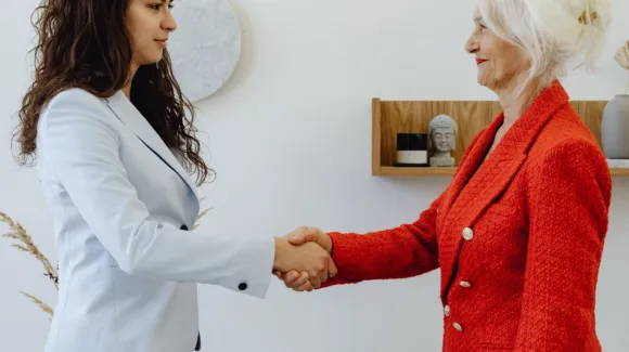 Two women in business attire are shaking hands in an office setting, standing in front of a shelf with a Buddha statue and decorative items.