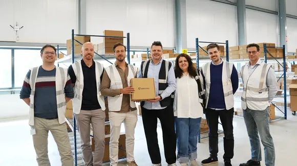 Seven people wearing safety vests stand in a warehouse with shelving and boxes, one person in the center holding a cardboard box and smiling at the camera.