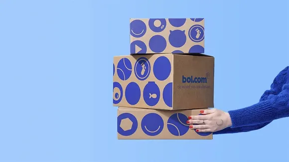 A hand holding a stack of three brown bol.com boxes with blue circular icons on a light blue background.