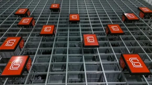 A warehouse automation system features red robotic units moving along a grid structure above storage bins.