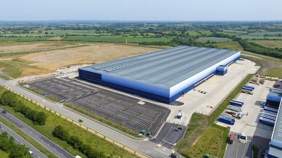 A large distribution warehouse with a mostly empty parking lot, surrounded by open fields and roads, viewed from above during daytime.