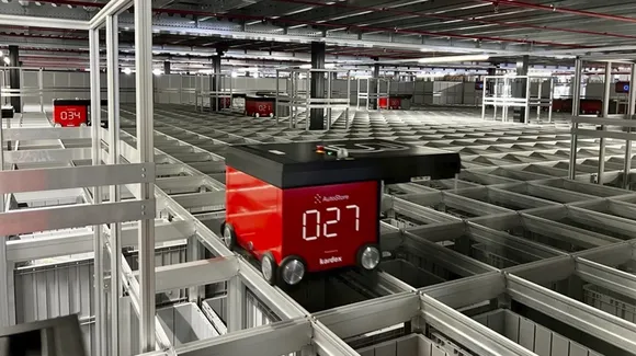 A red automated robot moves on tracks above a grid of storage bins in a warehouse, indicating the number 027 on its display.