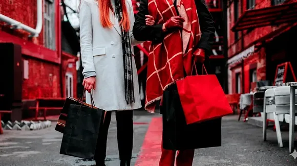 Two people standing on a city street holding multiple shopping bags, dressed in coats and scarves. Only their bodies are visible, not their faces.