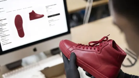 A person in black gloves holds a red leather high-top sneaker in front of a computer screen displaying a website with images of the shoe.