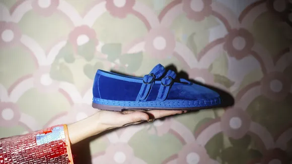 A person holds a blue slip-on shoe with two buckle straps against a patterned floral wallpaper background.