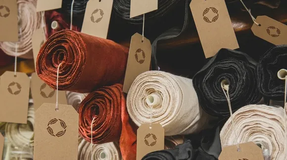 Rolls of fabric in various colors, including red, white, and black, stacked with brown tags featuring a circular arrow symbol attached to each roll.