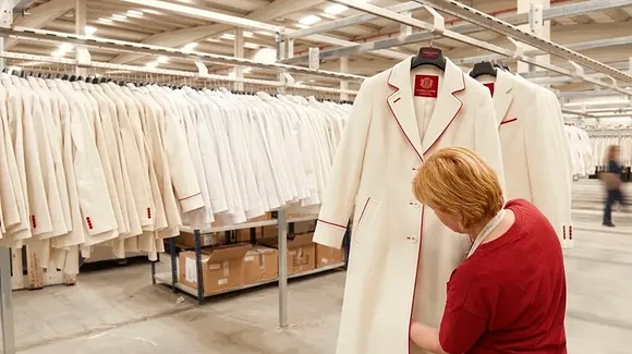 A person inspects a white coat with red trim in a warehouse filled with similar coats hanging on racks.