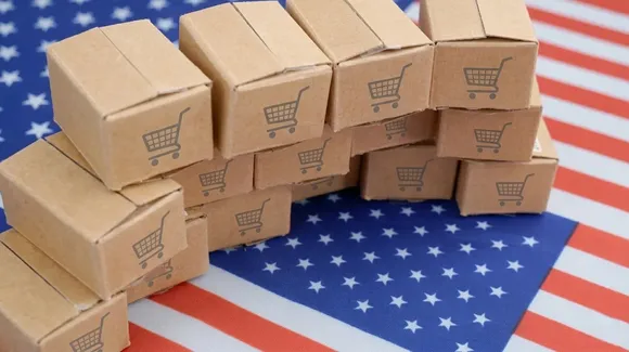 Stacked cardboard boxes with shopping cart symbols are arranged on a United States flag background.